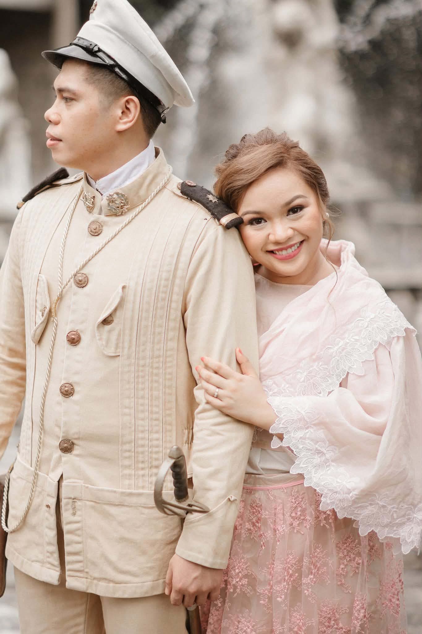 A couple in Filipiniana attire before a stone fountain — he in a cream uniform with a peaked cap and sabre, she in a blush baro't saya with an embroidered lace panuelo